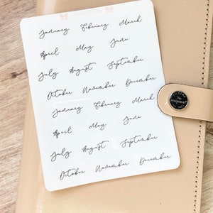 May include: A white sticker sheet with the months of the year written in elegant black cursive script. The sticker sheet is placed on a beige leather planner. The planner has a silver button that says "FILOFAX the original".