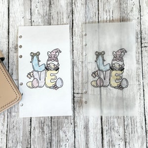 May include: Two planner inserts with the word "LOVE" in pastel colors, featuring a gnome wearing bunny ears. The inserts are decorated with Easter eggs and flowers. One insert is printed on white paper, the other is translucent.