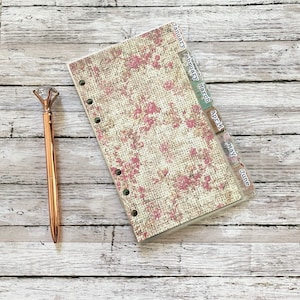 May include: A rose gold pen with a diamond-shaped top rests beside a planner. The planner features a floral design with pink flowers on a beige background. Tabs indicate the months January through June.