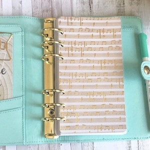 May include: A light teal binder with gold rings, containing a notepad printed with gold musical notes on a white backdrop. A gold paperclip and a pen are also visible. The binder is open, showing the inside.