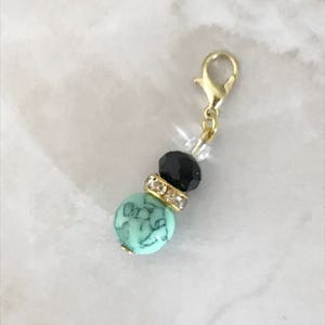 May include: A gold-colored lobster clasp charm with a turquoise bead, a black bead, and a clear bead. The turquoise bead has a marbled pattern. A gold-colored band with clear rhinestones separates the turquoise and black beads.