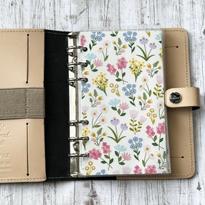 Op de afbeelding: Open beige leren planner met een bloemig patroon inlegvel. Het inlegvel heeft een witte achtergrond met roze, gele en blauwe bloemen. De planner heeft een zilveren ringband en een beige elastische sluiting.