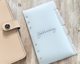May include: A translucent white planner insert labelled "January" in cursive script. The insert has tabbed dividers and is designed for a ring-bound planner. A beige leather planner is partially visible.