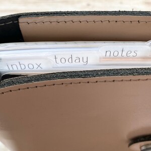 May include: A tan leather planner with a black interior and silver metal rings. The planner is open, revealing labeled dividers for "inbox", "today", "notes", and "finances". The planner is designed for organization and planning.