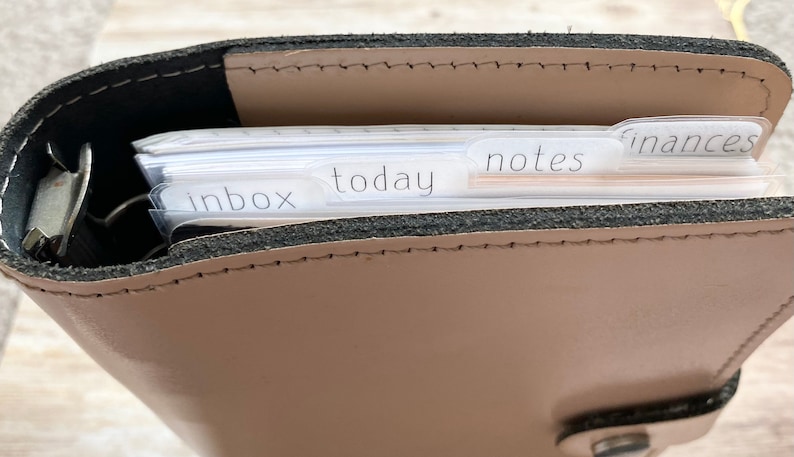 May include: A tan leather planner with a black interior and silver metal rings. The planner is open, revealing labeled dividers for "inbox", "today", "notes", and "finances". The planner is designed for organization and planning.