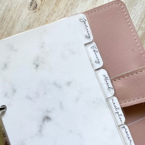 May include: A planner with clear dividers labeled January, February, March, meal plan, and groceries. The planner has a white marble design on the pages and a pink cover. The dividers have handwritten-style text.