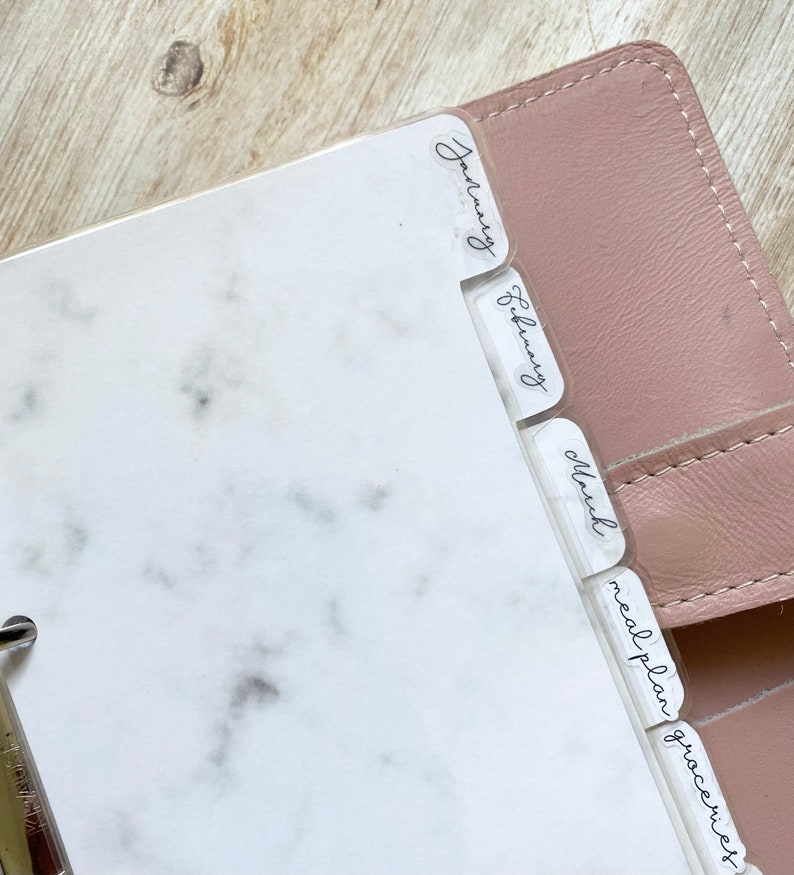 May include: A planner with clear dividers labeled January, February, March, meal plan, and groceries. The planner has a white marble design on the pages and a pink cover. The dividers have handwritten-style text.