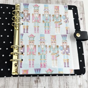 May include: A black and gold binder with a polka dot cover, open to reveal a page with a repeating pattern of colorful nutcracker illustrations. The nutcrackers are in shades of pink, blue, and gold, with various accessories.