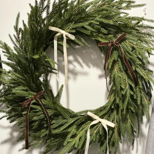 May include: A circular evergreen wreath with a full, natural look. The wreath features four decorative bows, two in cream and two in a deep brown velvet. A festive decoration for the home.