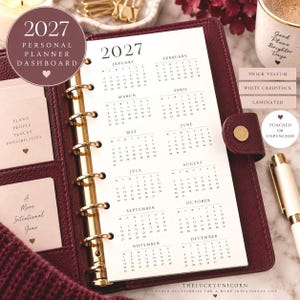 May include: A 2027 personal planner dashboard insert featuring a full-year calendar grid on white paper. The planner is held in a dark burgundy leather binder with gold rings. Text includes 2027, months from January to December, and inspirational phrases.