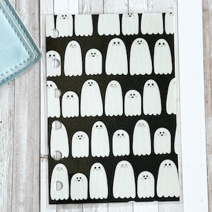 May include: A clear, plastic planner insert with a repeating pattern of white cartoon ghosts on a black background. The ghosts have simple, smiling faces. The insert has three ring binder holes.