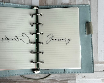 May include: A pale blue leather planner with lined paper pages. The word "January" is written in black script on a clear page divider. The word "Calendar" is also visible. The planner is open, revealing the metal ring binder.