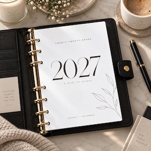 May include: A black leather binder with gold rings containing a 2027 planner insert. The white cover page features the text TWENTY TWENTY-SEVEN, 2027, a year to create, and JANUARY - DECEMBER, accompanied by a delicate line drawing of leaves.