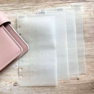 May include: A pink leather planner with clear, translucent dividers labeled "inbox", "today", "notes", and "finances". The dividers are designed for organization and are visible against a light wooden surface.