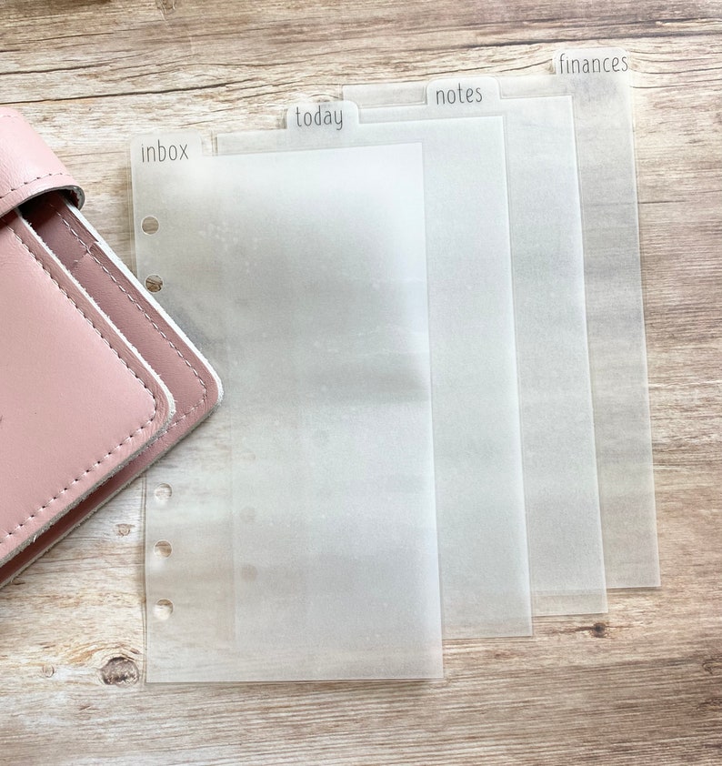 May include: A pink leather planner with clear, translucent dividers labeled "inbox", "today", "notes", and "finances". The dividers are designed for organization and are visible against a light wooden surface.