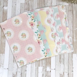 May include: A set of planner dividers with floral designs and monthly labels. The dividers feature a variety of pastel colors, including pink, yellow, and blue, with floral patterns and decorative circles. Each tab is labeled with a month.