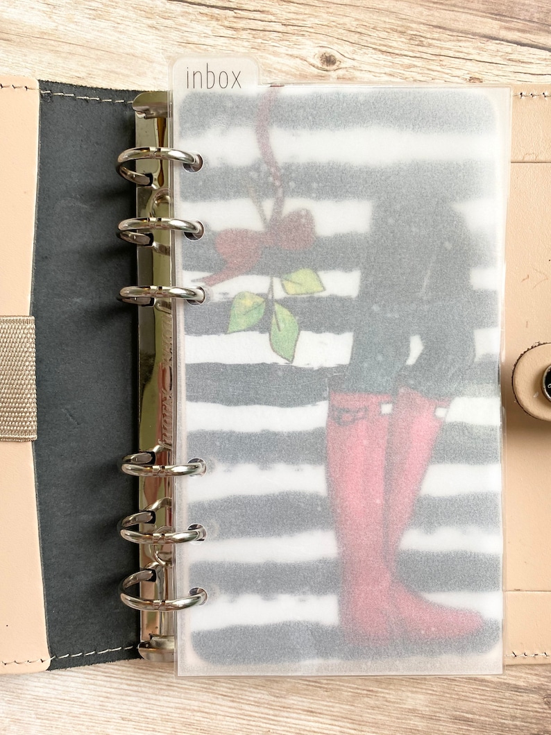 May include: A planner insert with a black and white striped background and a graphic of red rain boots. The insert is labeled "inbox" at the top and is held in a ring binder.