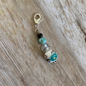 Puede incluir: Un charm de cuentas con un cierre plateado. El charm presenta una cuenta negra, una cuenta turquesa, una cuenta facetada transparente y una cuenta floral blanca con detalles azules. Para joyería o accesorios.