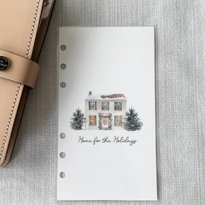 May include: A planner insert with a watercolor illustration of a house decorated for the holidays. The image features a white house with green wreaths and two small Christmas trees. The text "Home for the Holidays" is written below the house.