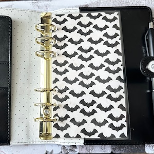 Peut inclure: Agenda en cuir noir avec des anneaux dorés, un stylo noir et un insert à motifs. L'insert présente un motif répété de chauves-souris noires sur fond blanc. L'agenda est ouvert, révélant l'intérieur avec une doublure à pois.