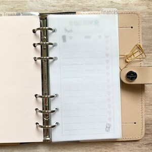 May include: A beige leather planner with a silver binder mechanism. A clear page with the words "Bills to Pay" and a list of dotted lines is visible. A gold paper clip and a "finances" tab are attached.