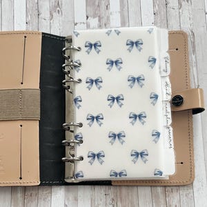May include: A beige leather planner with a black interior, open to reveal a section with a white cover patterned with blue bows. The planner has labeled dividers for February, March, meal plan, and groceries.