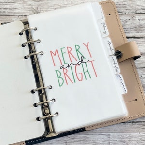 May include: Open planner with a white page featuring the words "MERRY and BRIGHT" in red and green. The planner has labeled dividers for months and categories. The planner is tan with silver rings.