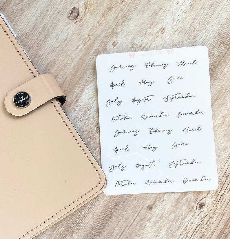 May include: A sheet of planner stickers with the months of the year written in elegant black script. The stickers are on a white background, next to a beige leather planner. The planner has a silver button that says "FILOFAX the original".