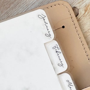 May include: Close-up of a planner with clear month dividers labeled January, February, and March. The planner is encased in a light tan leather cover with visible stitching and a small metal accent.