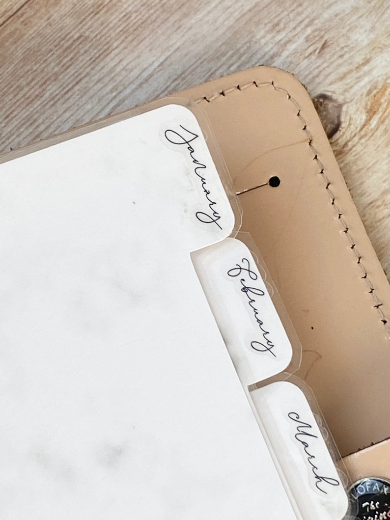 May include: Close-up of a planner with clear month dividers labeled January, February, and March. The planner is encased in a light tan leather cover with visible stitching and a small metal accent.