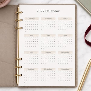 May include: A 2027 calendar page inserted into a beige ring-bound planner. The white page features twelve monthly grids arranged in three columns, each with a light beige checkered header. The gold-toned metal rings secure the paper in place.