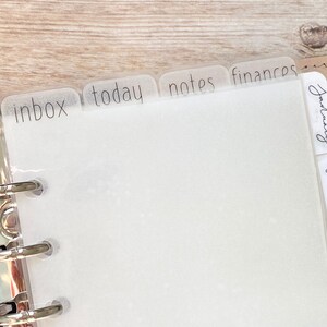 May include: A close-up of a planner with translucent dividers labeled "inbox", "today", "notes", and "finances". The planner is open, revealing blank white pages. The dividers are attached to a silver binder.
