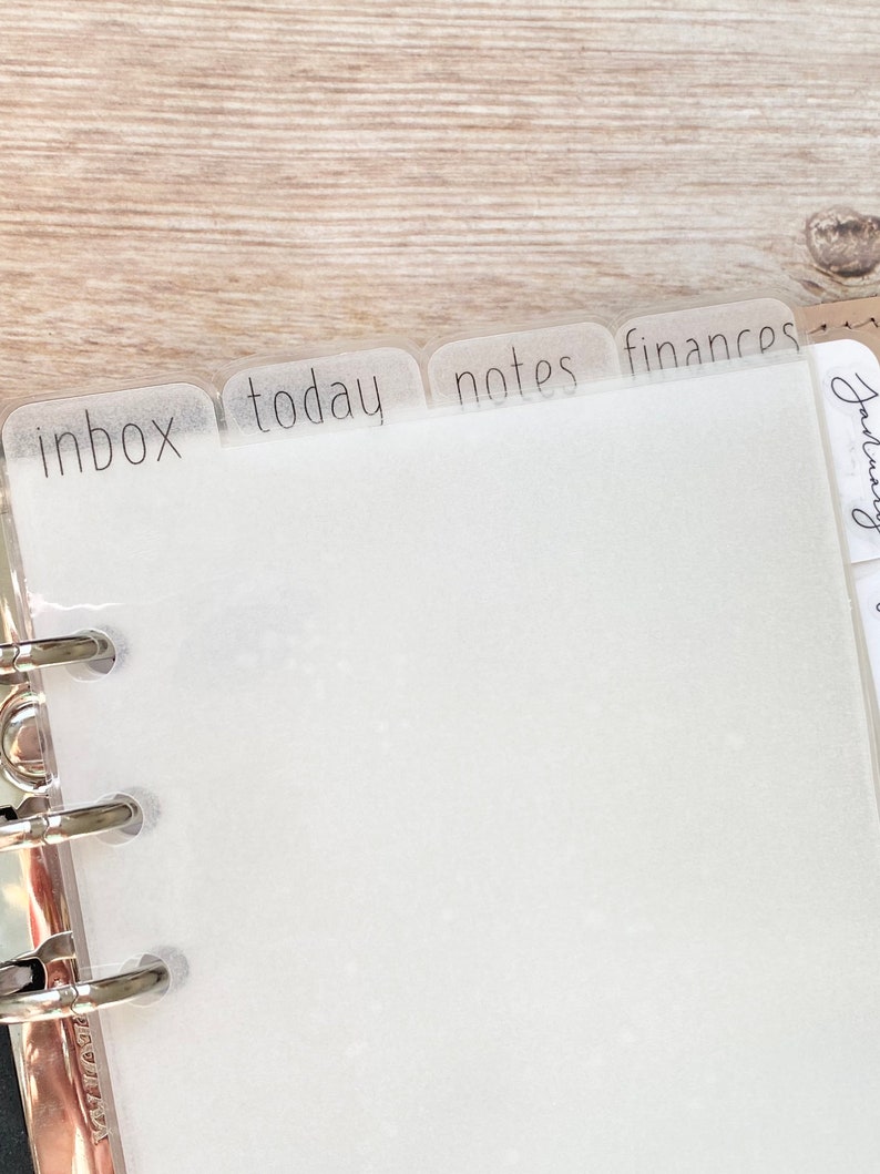 May include: A close-up of a planner with translucent dividers labeled "inbox", "today", "notes", and "finances". The planner is open, revealing blank white pages. The dividers are attached to a silver binder.