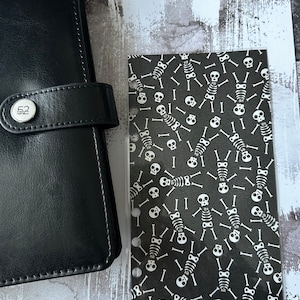 Könnte beinhalten: Schwarzer Lederplaner mit silbernem Verschluss und einem dekorativen Einsatz mit einem sich wiederholenden Muster aus weißen Skeletten auf schwarzem Hintergrund. Der Einsatz hat mehrere Löcher zum Binden.