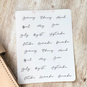 May include: A white sticker sheet with the months of the year written in elegant black cursive script. The months are arranged in rows, with two sets of each month. The sticker sheet is on a light wooden surface.