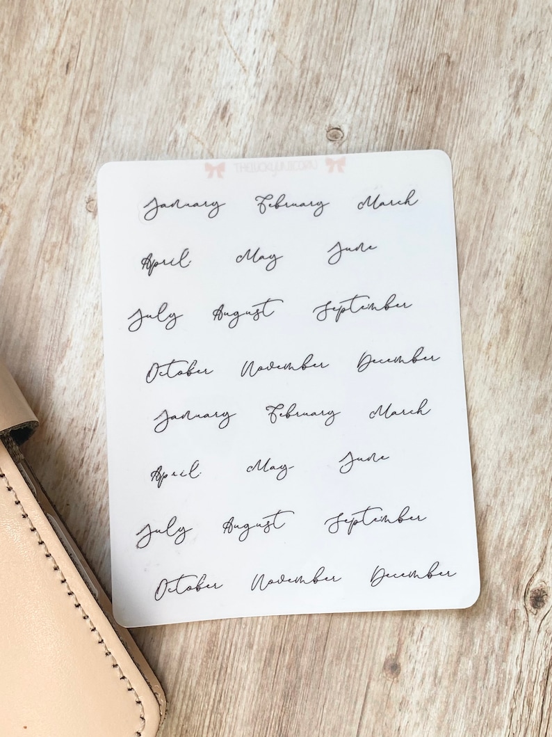 May include: A white sticker sheet with the months of the year written in elegant black cursive script. The months are arranged in rows, with two sets of each month. The sticker sheet is on a light wooden surface.