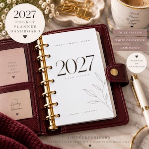 May include: A 2027 pocket planner dashboard featuring white cardstock with black text reading 2027, a year to create, and a line-art leaf illustration. The planner is held in a dark red leather binder with gold rings and includes small cards.