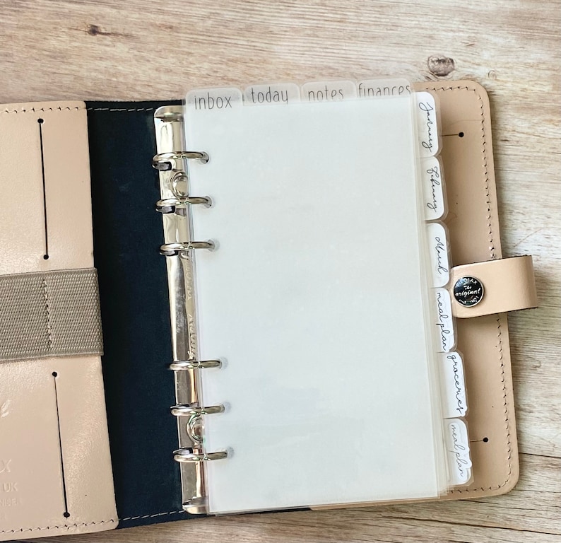 May include: Open leather planner with a silver binder mechanism. The planner has labeled dividers for "inbox", "today", "notes", and "finances". Additional dividers are labeled with months and other categories. The planner is open to blank white pages.