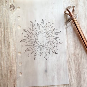 May include: A translucent planner insert featuring a celestial design with a sun and crescent moon illustration. The design is in black ink and includes stars and decorative elements. A rose gold pen with a diamond-shaped top rests on the wooden surface.