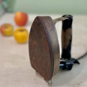May include: A vintage, rusty brown flat iron with a black handle and silver metal support. The iron is positioned on a light-colored surface, with three apples in the background. The apples are red and yellow.