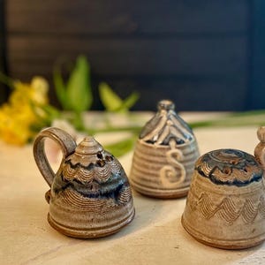 May include: Three handcrafted ceramic shakers with a speckled beige glaze and blue accents. Two shakers feature side handles and perforated tops, while the central shaker is bell-shaped with a prominent letter S embossed on the front surface.