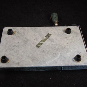 May include: A gray slate cheese board with a metal wire cheese cutter and a green handle. The board has four black rubber feet on the bottom.