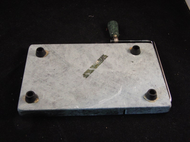 May include: A gray slate cheese board with a metal wire cheese cutter and a green handle. The board has four black rubber feet on the bottom.
