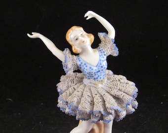 May include: A porcelain figurine of a ballerina wearing a blue and white polka dot dress with a lace overlay. The ballerina is posed in a dance move with one arm raised above her head.