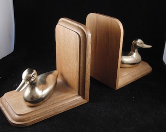 May include: A pair of wooden bookends with gold duck figurines. The bookends are made of a light brown wood with a smooth finish. The ducks are in a sitting position with their heads turned to the side.