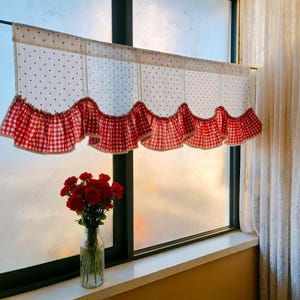 May include: A window valance with a white background and red polka dots and hearts. The bottom edge has a ruffled trim in a red and white gingham pattern. A bouquet of red roses sits in a clear glass vase on the windowsill.