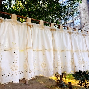 May include: White lace curtains with a scalloped edge and floral cutouts. The curtains are hung on a wooden rod with decorative bows. The curtains are made of a soft, flowing fabric, and are suitable for a window treatment.