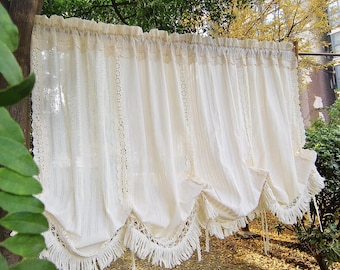 May include: Cream-colored curtain with lace trim and a fringed bottom edge. The curtain is made of a soft, textured fabric and is gathered at the top. The curtain is hanging outdoors, with greenery in the foreground and background.