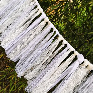 May include: A white lace garland featuring diverse lace designs. The garland is composed of numerous white lace strips, producing a layered appearance. The garland is set against a backdrop of green foliage.
