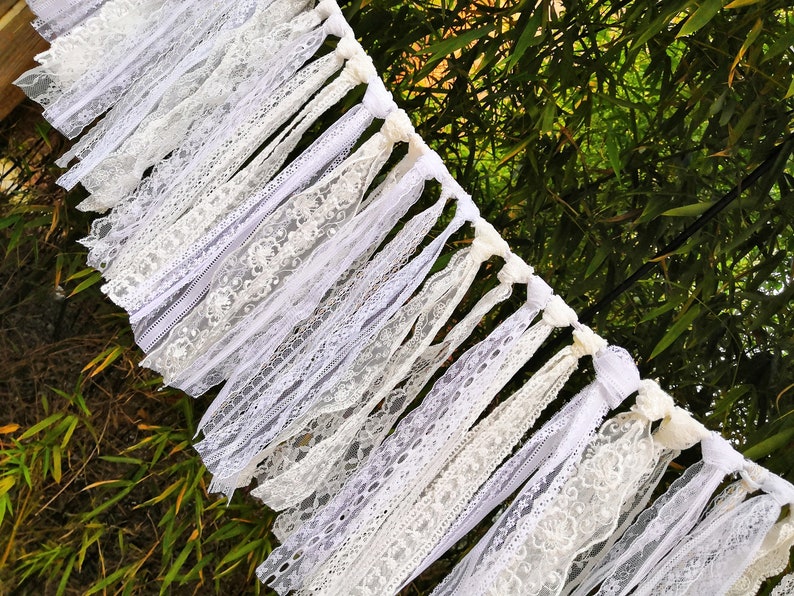 May include: A white lace garland featuring diverse lace designs. The garland is composed of numerous white lace strips, producing a layered appearance. The garland is set against a backdrop of green foliage.